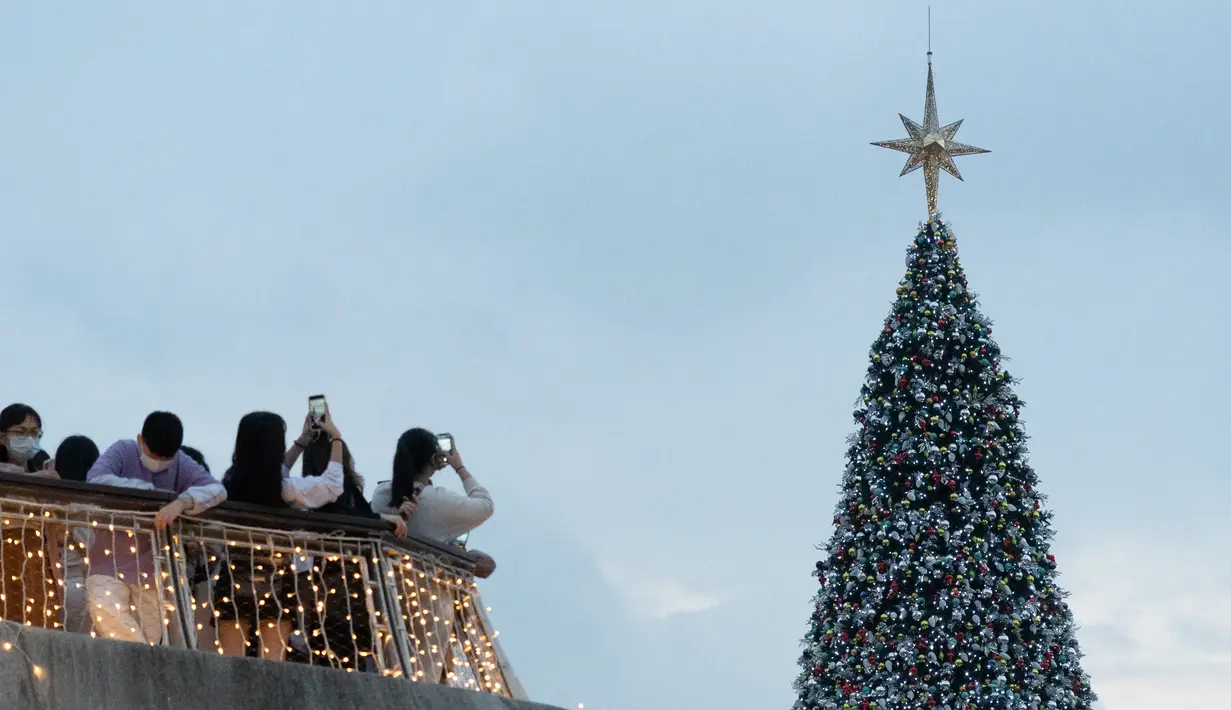 Orang-orang mengambil foto pohon Natal raksasa selama Natal di distrik budaya West Kowloon di Hong Kong (25/12/2021). Pohon Natal raksasa didekorasi secara glamor dengan karangan bunga dan ornamen, berkilauan dengan lampu meriah yang cerah. (AFP/Bertha Wang)