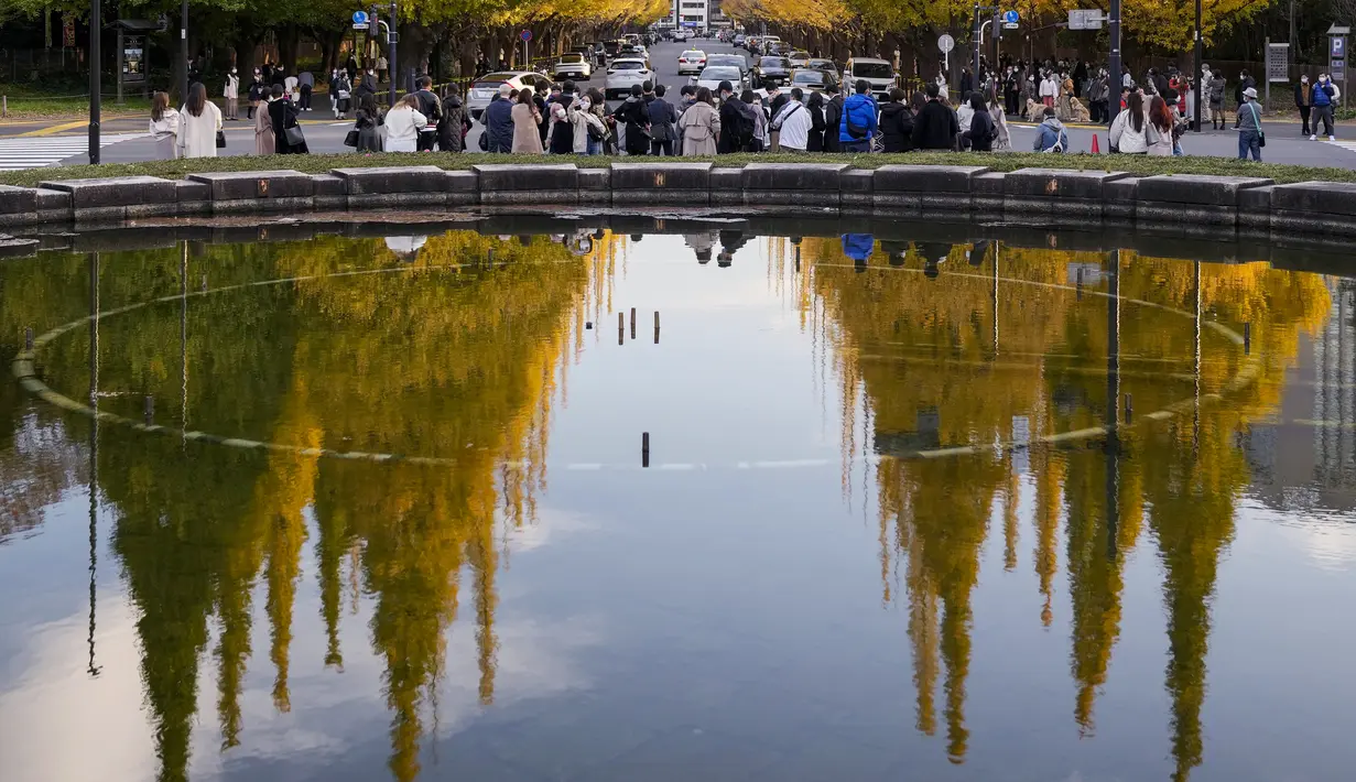 Orang-orang yang memakai masker pelindung untuk membantu mengekang penyebaran virus corona berjalan melalui deretan pohon ginkgo saat daun kuning cerah tercermin di permukaan air mancur  di Tokyo (29/11/2021). (AP Phot/Kiichiro Sato)