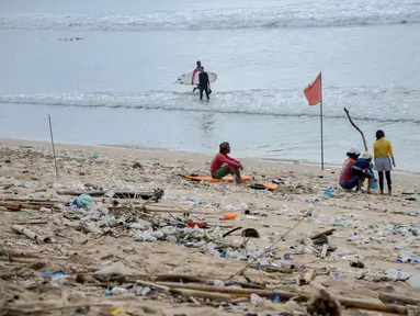 Orang-orang duduk di antara sampah kiriman yang terdampar memenuhi pesisir pantai Kuta, Bali, Kamis (31/12/2020). Menjelang pergantian tahun baru, Pantai Kuta hanya terlihat beberapa wisatawan, namun tumpukan sampah kiriman tersebar di sepanjang bibir pantai. (SONNY TUMBELAKA / AFP)