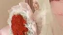 Larissa tampil cantik  mengenakan busana pengantin putih-putih.  Perempuan seorang anak itu tampak tersenyum bahagia duduk sambil memegang buket bunga mawar merah berukuran besar. [Instagram/larissachou]