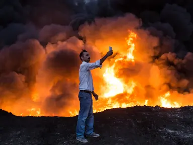 Seorang pria berselfie di depan kobaran api kilang minyak saat operasi perebutan kota Mosul oleh Pasukan Irak dari tangan ISIS, di Qayyarah, Mosul (19/10). Militer Irak telah berhasil menguasai beberapa wilayah ISIS di Mosul. (AFP Photo/YASIN Akgul)