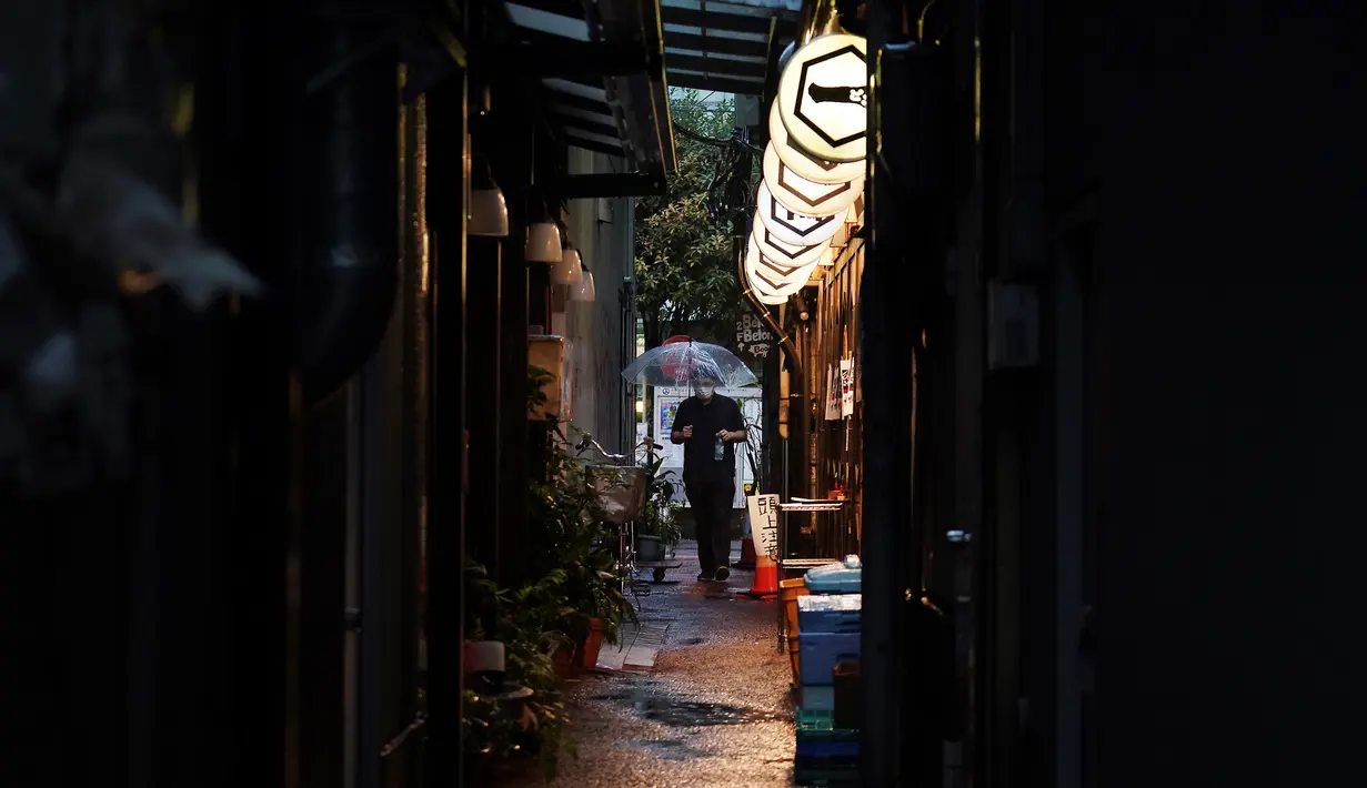 Seorang pria yang mengenakan masker untuk membantu mencegah penyebaran COVID-19 berjalan melewati gang sempit saat hujan di Tokyo, Jepang, Selasa (30/6/2020). Sebagian besar kegiatan sosial dan bisnis di Jepang telah kembali dimulai. (AP Photo/Eugene Hoshiko)
