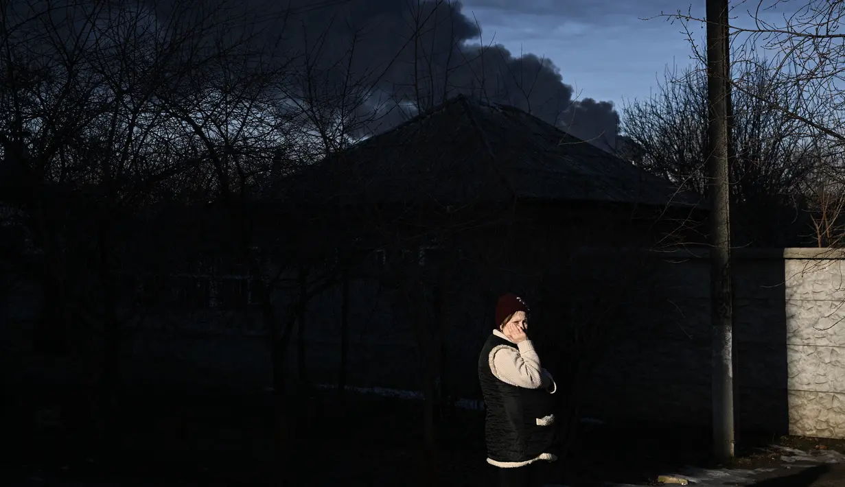 Seorang wanita berdiri di jalan saat asap hitam mengepul dari bandara militer di Chuguyev, dekat Kharkiv, 24 Februari 2022. Presiden Rusia Vladimir Putin mengumumkan operasi militer di Ukraina. (Aris Messinis/AFP)