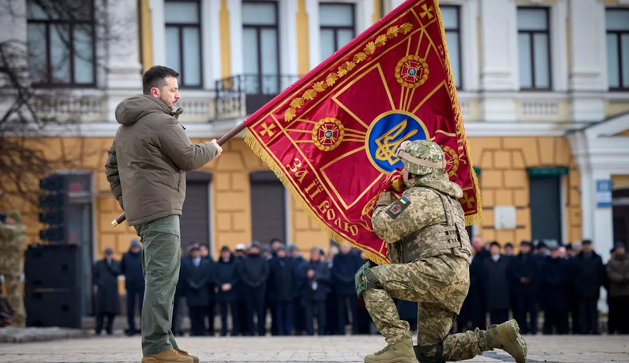 Presiden Ukraina Volodymyr Zelenskyy (kiri) memegang bendera unit militer saat seorang perwira menciumnya dalam acara peringatan satu tahun perang Rusia-Ukraina di Kyiv, Ukraina, Jumat, 24 Februari 2023. Volodymyr Zelenskyy berjanji akan terus berjuang untuk mencapai kemenangan saat memperingati satu tahun invasi Rusia yang disebutnya sebagai “hari terpanjang dalam hidup kita.” (Kantor Pers Kepresidenan Ukraina via AP)