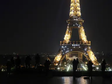 Awak media berdiri di depan Menara Eiffel dengan tulisan 'Merci" (terima kasih) yang terpampang di Paris, Jumat (27/3/2020). Menara Eiffel menampilkan pesan dukungan dan terima kasih kepada tenaga kesehatan di Prancis yang berada di garda depan memerangi pandemi COVID-19.  (AP/Thibault Camus)