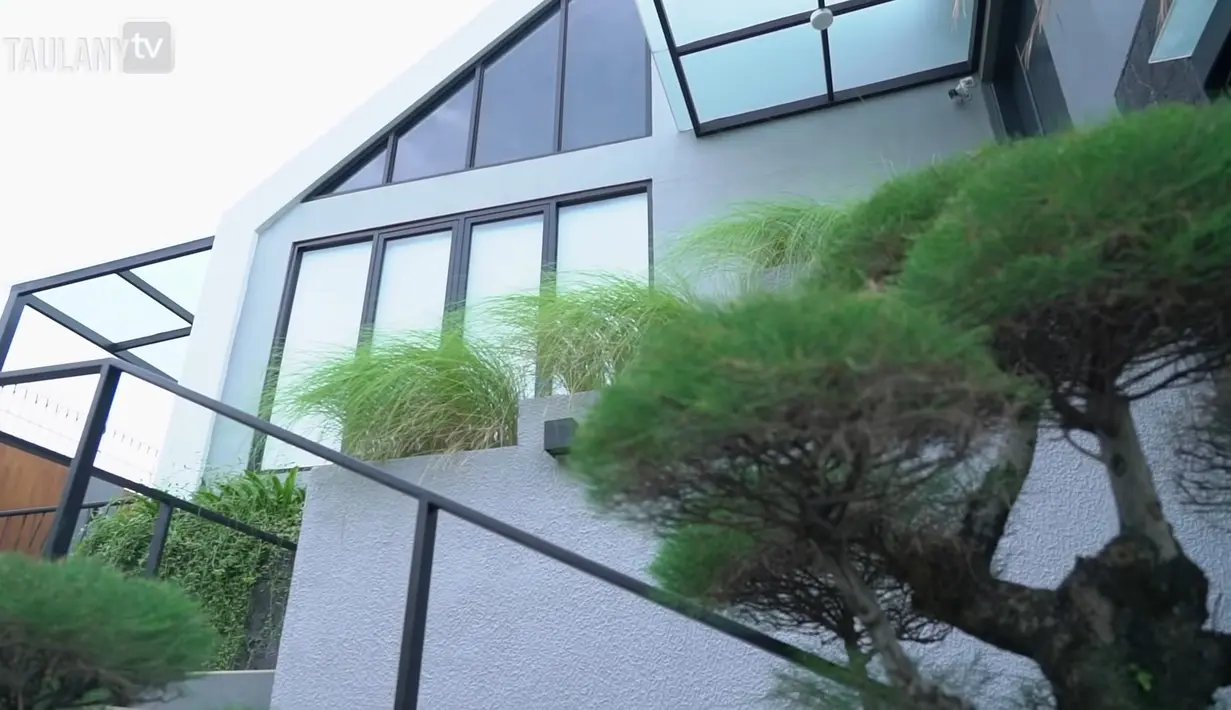 Bagian luar rumah memiliki daya tarik dengan taman terbuka yang asri. Bangunan dipenuhi kaca tinggi yang mempercantik tampilan rumah. (YouTube Taulany TV)