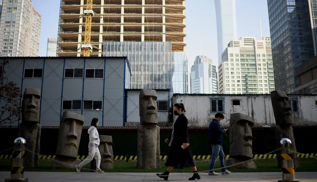 Orang-orang yang memakai masker berjalan melewati replika patung raksasa 'Moai' Pulau Paskah di Beijing pada Rabu (4/11/2020). Pulau Paskah yang berada di Negara Chile terkenal oleh patung kepala yang dipahat dari batu. (Photo by Noel Celis / AFP)