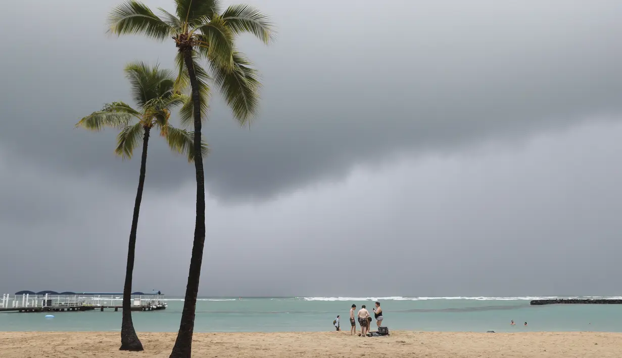 <p>Para pengunjung pantai terlihat dengan latar belakang awan badai, di Pantai Waikiki, Honolulu (6/12/2021). Badai kuat yang membawa angin kencang dan hujan yang sangat deras membanjiri jalan-jalan dan meruntuhkan kabel listrik dan cabang-cabang pohon di seluruh Hawaii. (AP Photo/Marco Garcia)</p>