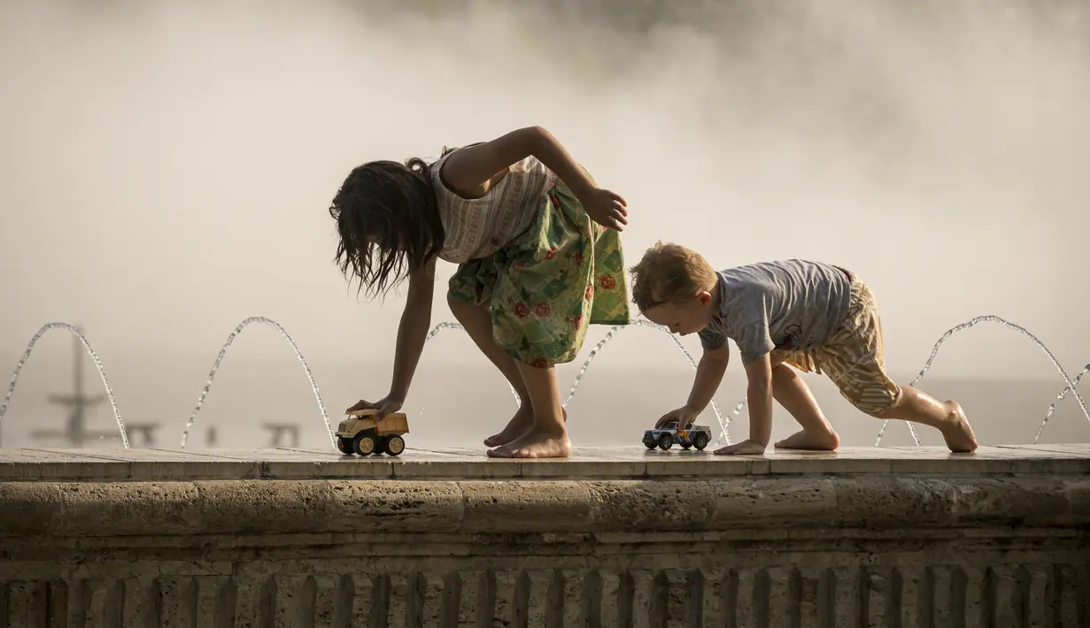 Anak-anak bermain dengan mobil mainan berlatar kabut dari air mancur umum di Bukares, Rumania, Kamis (13/7/2023). (AP Photo/Andreea Alexandru)