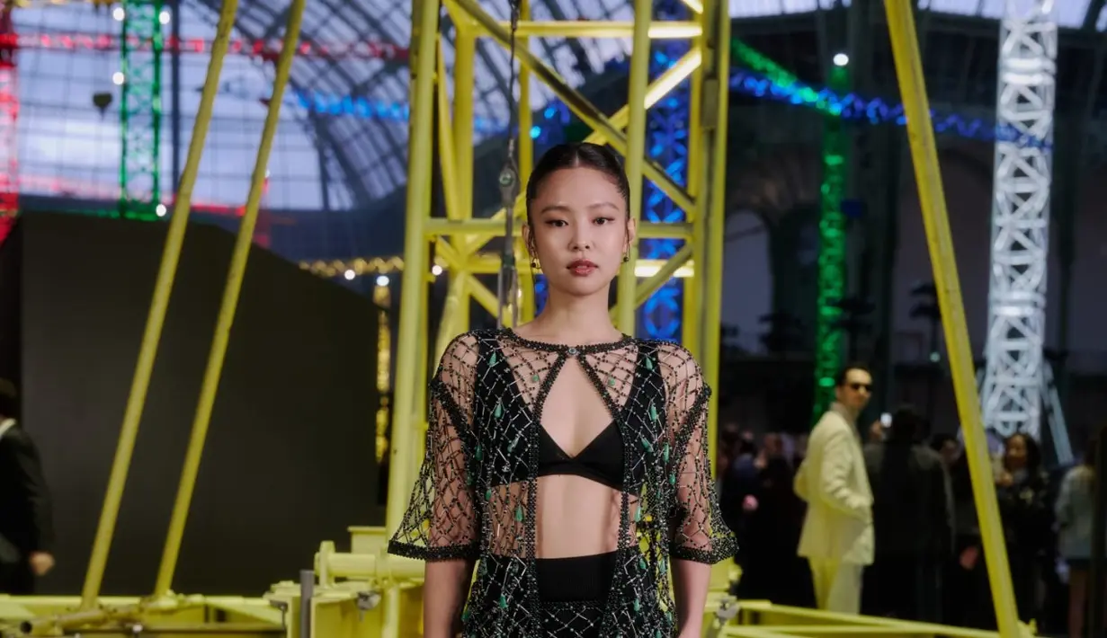 Sebagai global ambassador Chanel, Jennie BLACKPINK hadir dengan gaya yang jauh lebih edgy. Ia mengenakan set transparan berbahan mesh yang dihiasi bead dan kristal berkilau, dipadukan dengan bralette hitam dan high-waist shorts sebagai inner. Siluetnya memperlihatkan permainan layer yang sensual namun tetap sophisticated.