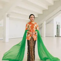 Menyanyikan lagu Kebaya bersama 5 Wanita, Andien Aisyah kenakan kebaya Janggan bermotif bunga warna oranye. Dipadukan dengan kain Batik cokelat dan selendang berwarna hijau. Rambutnya pun ditata bergaya sanggul tekuk dengan tusuk konde di bagian samping. [@andienaisyah].