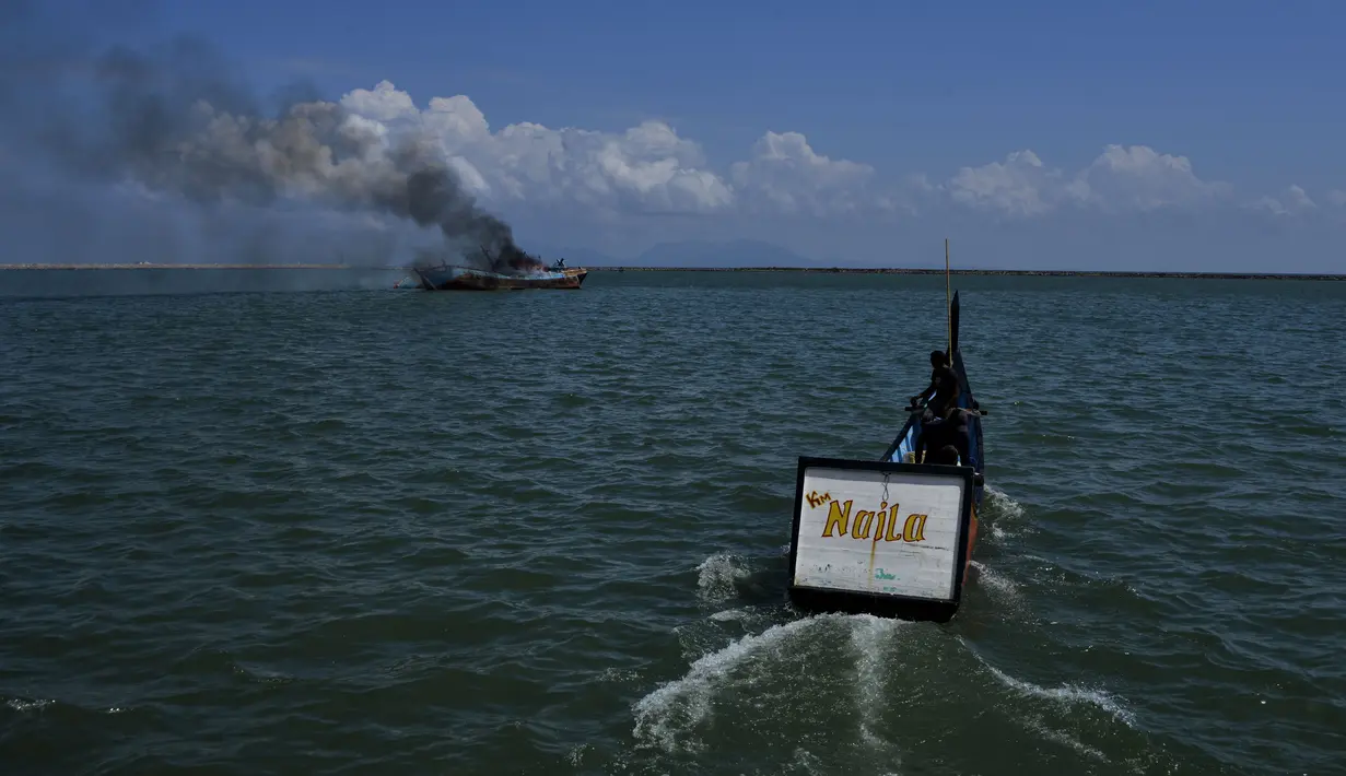 Seorang nelayan (kanan) berlayar melewati asap mengepul dari kapal asing ilegal asal Malaysia yang dibakar dan ditenggelamkan Kejaksaan Negeri Banda Aceh di perairan Pelabuhan Perikanan Samudera Kutaraja, Banda Aceh, Aceh, Kamis (18/3/2021). (AFP/Chaideer Mahyuddin)