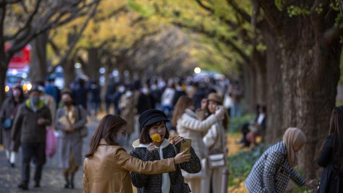 Orang-orang dengan mengenakan masker mengambil foto saat mereka berjalan melalui barisan pohon ginkgo saat pepohonan dan trotoar ditutupi dedaunan kuning cerah di sepanjang trotoar di Tokyo, Jepang pada 28 November 2020. (AP Photo/Kiichiro Sato)