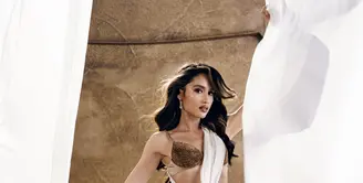 Cinta Laura tampil bak Dewi Athena dengan gaun putih berbahan chiffon yang dililitkan asimetris dan berpotongan cut-out di pinggang. Atasan berhias kristal emas serta bros bunga matahari dari batu kristal besar menjadi pusat perhatian, dipadukan sayap bulu emas yang megah dan heels gladiator emas bertali tinggi. [@claurakiehl]