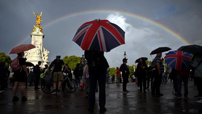 Seseorang dengan payung bertema bendera Union berlindung dari hujan di seberang Istana Buckingham, saat pelangi ganda muncul di atas langit London,  Kamis, 8 September 2022. Pelangi ganda muncul di atas kastil pada hari wafatnya Ratu Elizabeth II, selama beberapa menit dan kemudian menghilang begitu saja. (Photo by Daniel LEAL / AFP)
