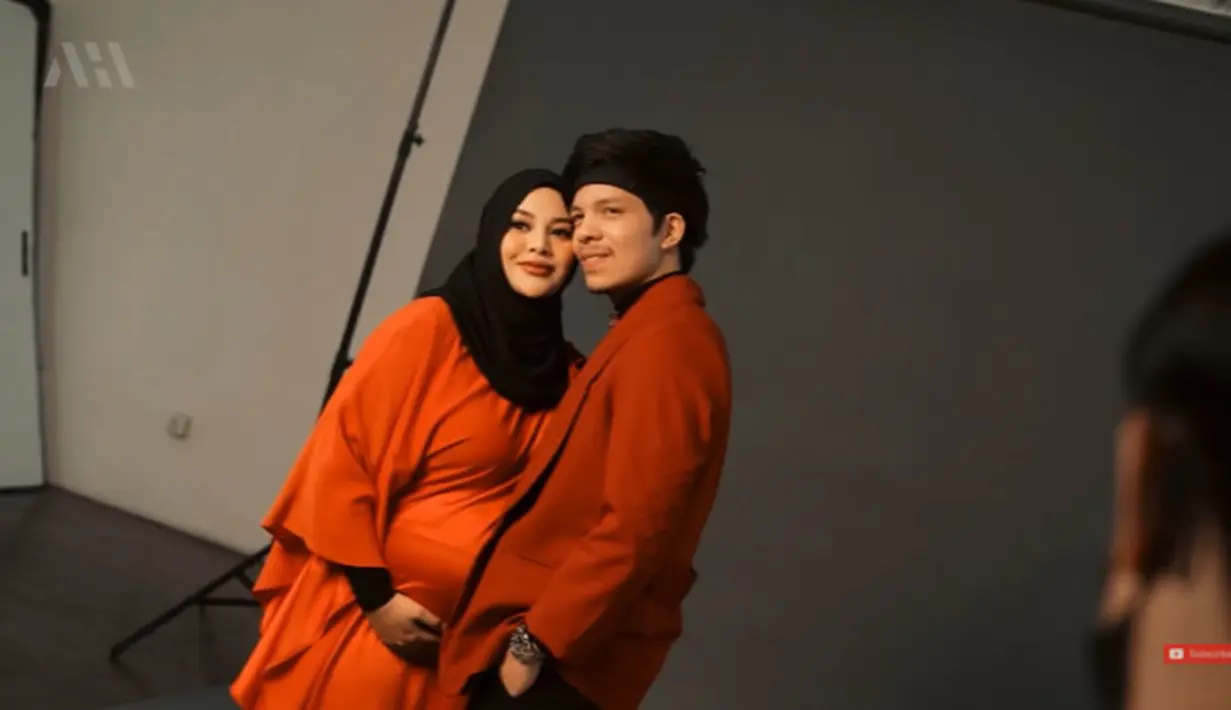 Aurel mengenakan dress satin berwarna oranye dengan hijab berwarna hitam. Sementara, Atta Halilintar mengenakan crew neck dan jas oranye. Tak ketinggalan headband andalan Atta juga ia kenakan. (YouTube/AH)