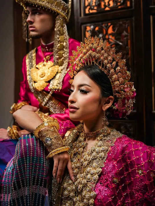 Atasan satin mengilap dipadukan dengan sarung bermotif kotak khas membuat tampilannya terlihat mencolok sekaligus berkelas. Ia juga mengenakan aksesori kepala dan selempang emas penuh detail yang memberi nuansa bangsawan tradisional. [@jennifercoppenreal20].