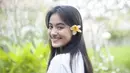 Mawar Eva memamerkan senyum manisnya ke kamera dengan bunga kamboja yang diselip pada telinganya. Mawar membagikan fotonya dengan background tanaman hijau yang terlihat buram. (Instagram/@mawar_eva)