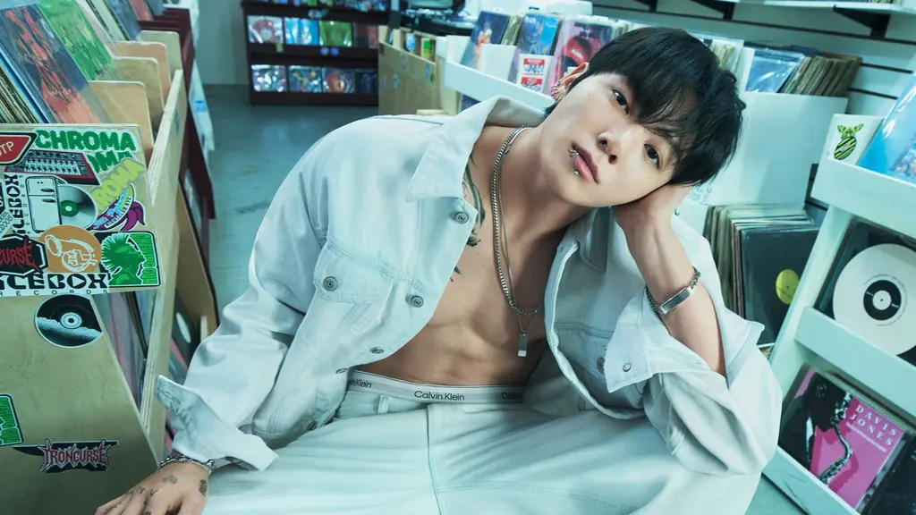 Dalam foto pertama, Jung Kook tampil berani dengan jaket denim putih yang dibiarkan terbuka tanpa inner, memperlihatkan torso atletisnya. Dipadukan dengan jeans senada berpotongan relaxed, gaya ini memancarkan maskulinitas raw yang tetap clean berkat palet monokrom. Pose santai di dalam toko piringan hitam memberi nuansa edgy sekaligus youthful.