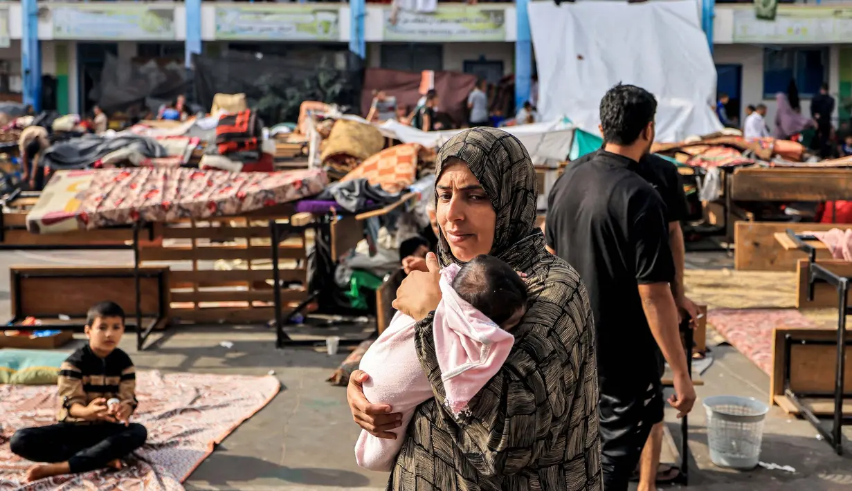 <p>Seorang wanita berjalan sambil menggendong bayi di taman bermain di sebuah sekolah yang dikelola oleh Badan Bantuan dan Pekerjaan PBB untuk Pengungsi Palestina (UNRWA) yang telah diubah menjadi tempat penampungan bagi para pengungsi Palestina di Khan Yunis di Jalur Gaza selatan pada 25 Oktober 2023. (Mahmud HAMS/AFP)</p>