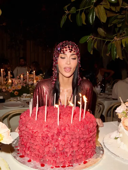 Kim meniup lilin pada kue raspberry segar yang menjulang, dengan lilin ramping berwarna pucat. Penutup kepala kristal merah dan gaun mesh metalik berkilau menjadi sorotan di tengah suasana makan malam dengan dekorasi bunga dan lilin [@kimkardashian]