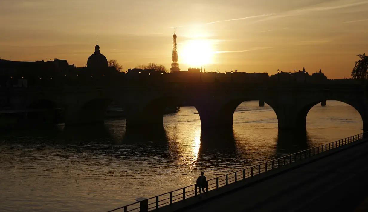 Seorang pria berjalan di tepi sungai Seine dengan latar belakang Menara Eiffel, saat matahari terbenam, di Paris (29/3/2021). Sungai ini membelah kota Paris menjadi dua bagian yang dalam bahasa Prancis disebut la rive droite ("tepi kanan") dan la rive gauche ("tepi kiri"). (AP Photo/Thibault Camus)