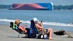 Rachel (kiri) dan Sarah Mitchell bermain ponsel saat berjemur dengan pemandangan kapal kargo Golden Ray yang terbalik di lepas pantai negara bagian Georgia, Amerika Serikat, Minggu (8/9/2019). Kapal tersebut dilaporkan memiliki 23 awak dan seorang nakhoda . (AP Photo/Terry Dickson)