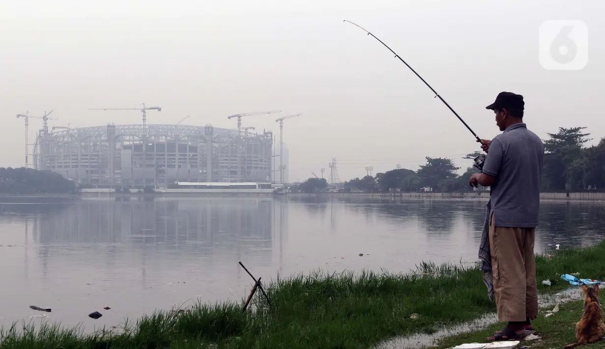 Warga memancing di pinggir Waduk Cincin yang berlatar pembangunan Jakarta International Stadium (JIS), Sabtu (19/6/2021). Pembangunan JIS sudah memasuki tahap pemasangan rangka atap dengan bentang mencapai 269 meter dan ditarget selesai Desember 2021. (Liputan6.com/Helmi Fithriansyah)