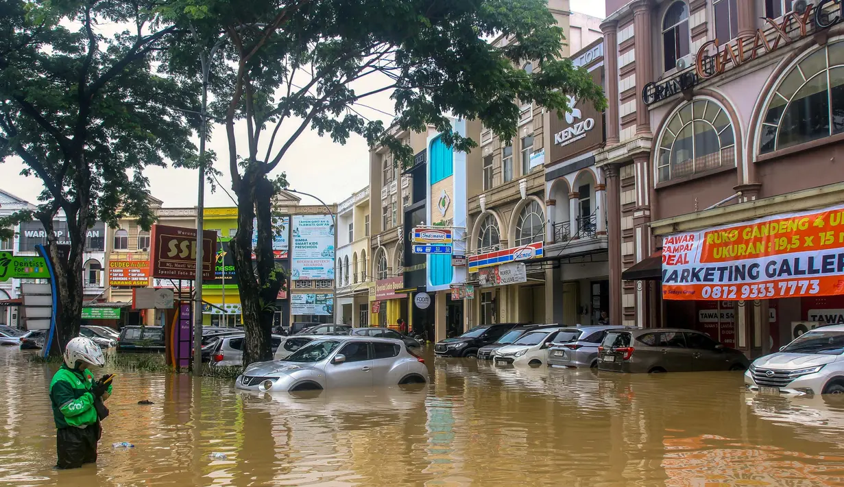 Seorang pengemudi ojek online menggunakan ponsel pintar sambil berdiri di tengah banjir yang merendam beberapa mobil di Kawasan Pertokoan Grand Galaxy di Bekasi, Jawa Barat pada Selasa 4 Maret 2025. (Robby Bibir Fathan Qorib/AFP)
