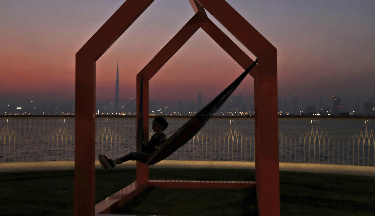 Seorang gadis berayun ketika matahari terbenam di belakang cakrawala kota dengan latar gedung tertinggi di dunia Burj Khalifa, di Dubai, Uni Emirat Arab (18/10/2019). (AP Photo/Kamran Jebreili)