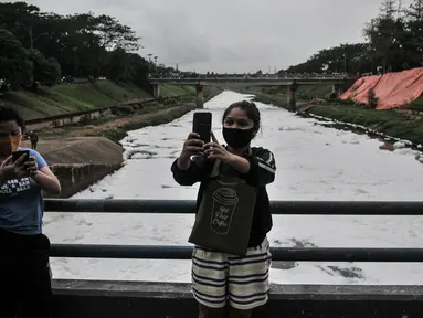 Warga berswafoto dengan latar belakang limbah busa yang memenuhi aliran Kanal Banjir Timur (KBT), Duren Sawit, Jakarta, Kamis (2/7/2020). Penampakan busa akibat limbah pabrik dan rumah tangga sejak dua minggu lalu itu menjadi tontonan warga meski menimbulkan bau tak sedap (merdeka.com/Iqbal Nugroho)