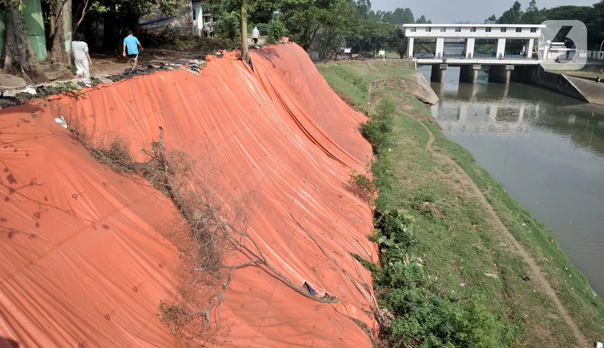 Warga melintasi lokasi tanah longsor yang ditutupi terpal besar di pinggir Kanal Banjir Timur, Jakarta, Kamis (18/6/2020). Warga setempat mengaku khawatir terjadi longsor susulan karena terpal dan karung pasir yang digunakan untuk menutupi lokasi longsor kian rapuh. (merdeka.com/Iqbal S. Nugroho)