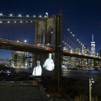 Gambar korban diproyeksikan pada Jembatan Brooklyn saat Hari Peringatan COVID-19 di Brooklyn, New York, Amerika Serikat, 14 Maret 2021. Kota New York yang mencatat kematian tertinggi akibat COVID-19 di AS, memberikan penghormatan yang emosional pada para korban yang meninggal. (Kena Betancur/AFP)