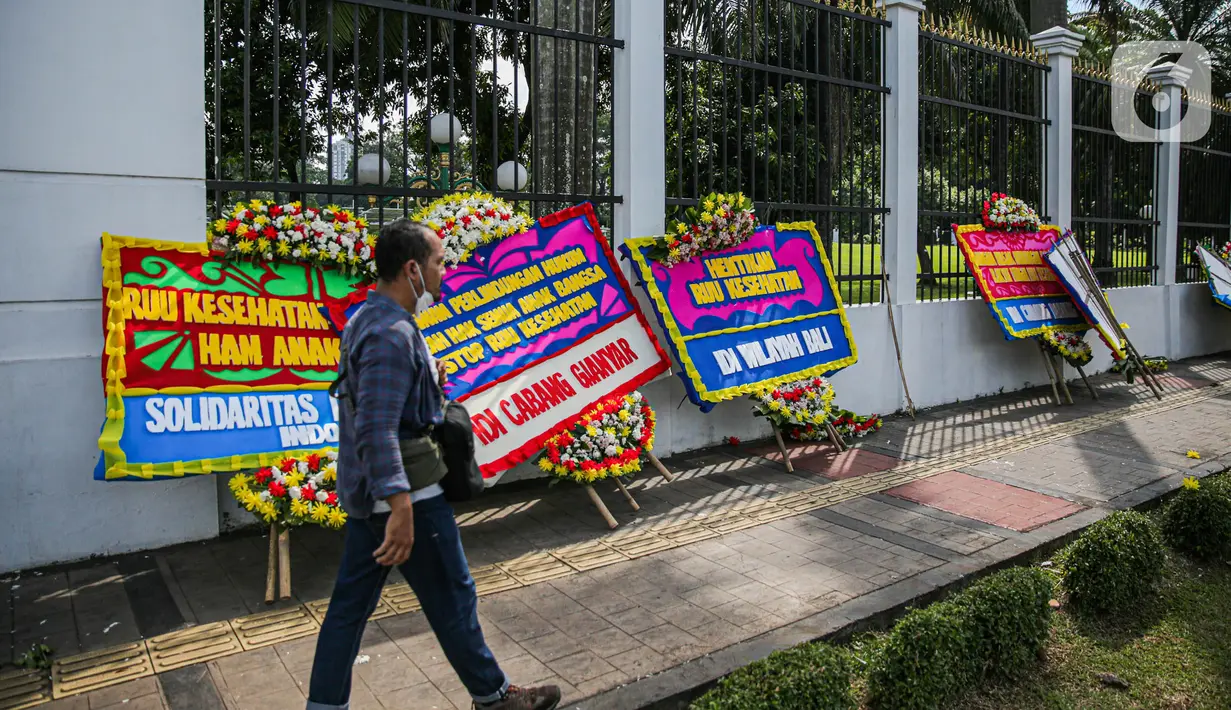 Seorang pria melintas di depan karangan bunga menolak RUU Kesehatan Omnibuslaw menghiasi depan gerbang Gedung DPR, Jakarta, Rabu (12/4/2023). (Liputan6.com/Faizal Fanani)