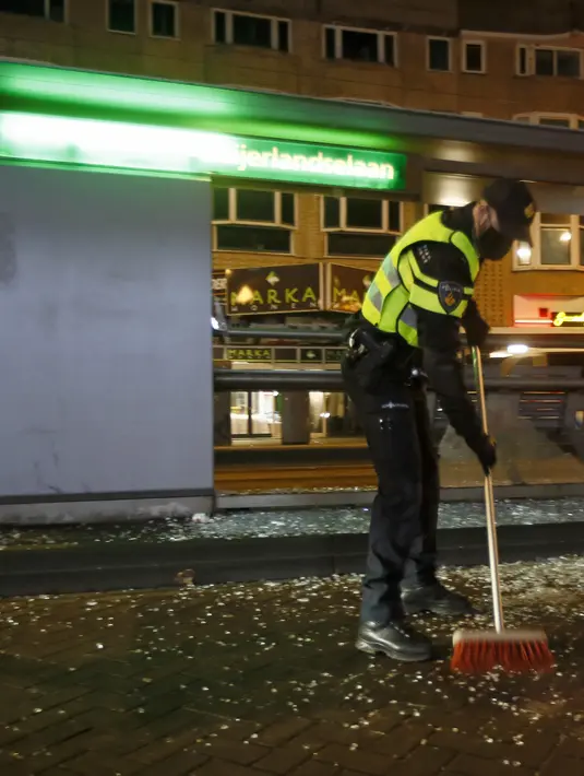 Seorang petugas polisi menyapu kaca dari halte bus yang dihancurkan dalam protes terhadap jam malam nasional di Rotterdam, Belanda, Senin (25/1/2021).  Bentrokan terjadi di beberapa lokasi di Belanda, termasuk di Amsterdam, akibat kebijakan jam malam untuk mencegah COVID-19. (AP Photo/Peter Dejong)