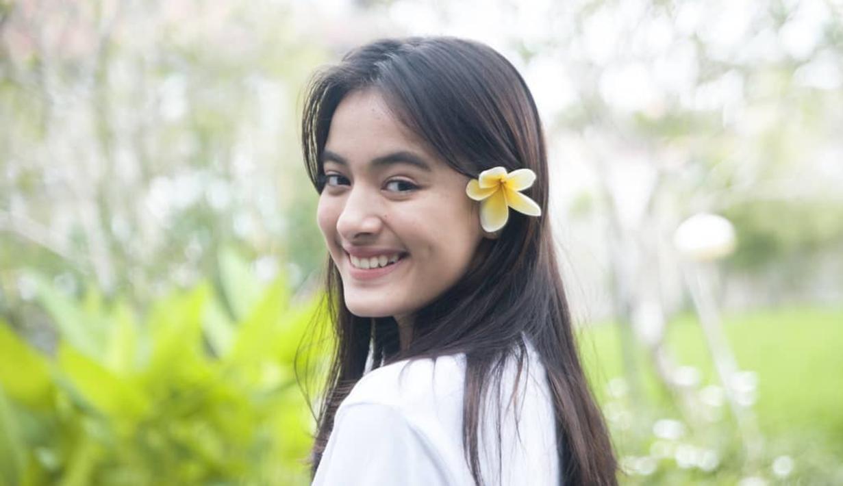 Mawar Eva memamerkan senyum manisnya ke kamera dengan bunga kamboja yang diselip pada telinganya. Mawar membagikan fotonya dengan background tanaman hijau yang terlihat buram. (Instagram/@mawar_eva)