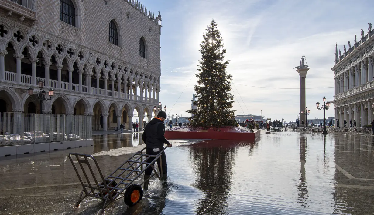 Seorang porter mengarungi St Mark's Square  yang dibanjiri air laut di samping pohon Natal di Venesia, Italia, Sabtu (4/12/2021). Air mencapai 99 sentimeter di atas permukaan laut dan bagian terendah kota tenggelam. (AP Photo/Luigi Costantini)