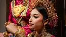 Atasan satin mengilap dipadukan dengan sarung bermotif kotak khas membuat tampilannya terlihat mencolok sekaligus berkelas. Ia juga mengenakan aksesori kepala dan selempang emas penuh detail yang memberi nuansa bangsawan tradisional. [@jennifercoppenreal20].