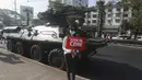 Seorang pengunjuk rasa anti-kudeta memegang poster saat berpose di depan kendaraan lapis baja yang dikerahkan di luar gedung Bank Sentral Myanmar di Yangon, Senin (15/2/2021). Pasukan militer Myanmar mulai menerjunkan kendaraan lapis baja di kota-kota besar dan memutus akses internet. (AP Photo)