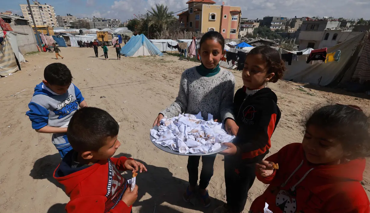Seorang gadis muda Palestina menjual permen kepada anak-anak di sebuah kamp di Rafah, Jalur Gaza selatan pada 7 Maret 2024. Perang dan pembatasan oleh Israel membuat warga Gaza dihantui sejumlah krisis akibat kelangkaan makanan, air, obat-obatan serta potensi penyebaran penyakit imbas lemahnya layanan medis. (MOHAMMED ABED / AFP)