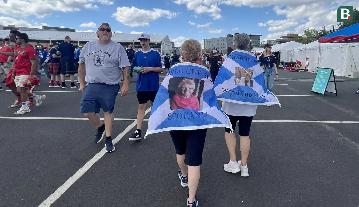 Suporter Skotlandia berjalan di area fanzone Gillette Stadium, Foxborough, Amerika Serikat, Jumat (19/6/2026), menjelang laga melawan Maroko pada Piala Dunia 2026. Mereka mengenakan jubah bergambar dan bendera Saltire sebagai bentuk dukungan sekaligus penghormatan kepada sesama anggota Tartan Army. (KLY Sports/Hery Kurniawan)