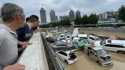 Orang-orang melihat mobil-mobil yang terendam banjir setelah hujan lebat melanda kota Zhengzhou di provinsi Henan, China tengah (21/7/2021). Luapan sungai menggenangi jalan-jalan dan membuat kendaraan terbawa arus setelah curah hujan 200 mm turun dalam satu jam. (AFP/STR)
