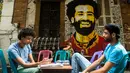 Dua pemuda duduk bersantai dengan latar belakang mural bintang Liverpool, Mohamed Salah di sebuah kedai kopi di pusat kota Kairo, 30 April 2018. Mural pemain timnas Mesir tersebut menghiasi sudut negara asalnya. (AFP Photo/KHALED DESOUKI)