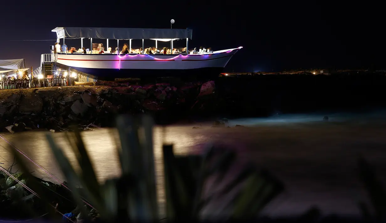 Warga Palestina menyantap hidangan sambil menikmati senja di sebuah restoran berbentuk perahu di tepi pantai Kota Gaza, Jalur Gaza, Rabu (2/8). Restoran tersebut berada di atas batu karang dengan hiasan lampu warna-warni. (MOHAMMED ABED / AFP)