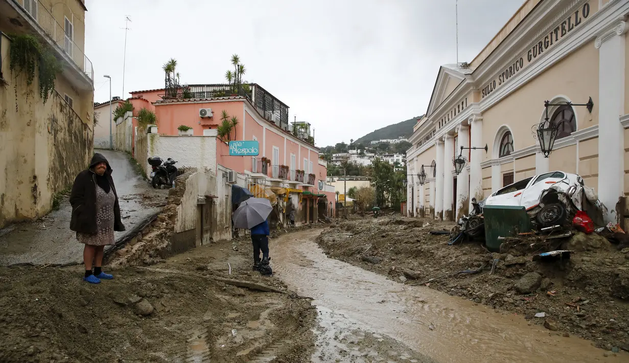 Orang-orang berjalan melalui lumpur di jalan yang banjir setelah hujan deras memicu tanah longsor yang meruntuhkan bangunan dan menyebabkan sebanyak 12 orang hilang, di Casamicciola, di pulau selatan Italia Ischia, Sabtu, 26 November 2022. Petugas pemadam kebakaran sedang melakukan upaya penyelamatan karena bala bantuan sedang dikirim dari Napoli terdekat, tetapi mengalami kesulitan dalam mencapai pulau baik dengan perahu motor atau helikopter karena cuaca. (AP Photo/Salvatore Laporta)