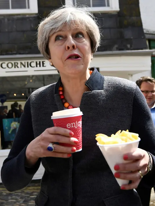 PM Inggris, Theresa May bereaksi sambil membawa keripik dan minuman saat berhenti untuk kampanye di Mevagissey, Cornwall, Selasa (2/5). Foto ini menjadi perbincangan karena Theresa May terlihat konyol saat mengemil. (Dylan Martinez/PA via AP)