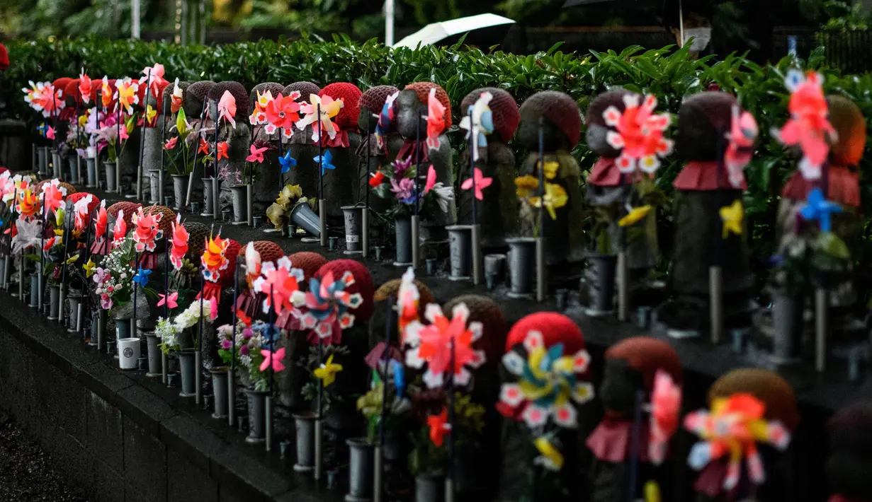 Patung-patung Jizo terlihat saat seorang pria dengan mengenakan payung melintas di Kuil Zojoji, Tokyo pada 22 Juni 2020. Jizo adalah pelindung dari anak-anak yang belum sempat lahir atau meninggal dalam kandungan dan saat masih bayi. (Photo by Philip FONG / AFP)