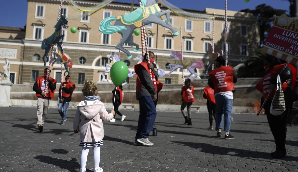 Seorang anak memegang balon saat melihat pekerja taman hiburan memegang komidi putar sementara mereka bergabung dengan pekerja sirkus dalam demonstrasi menuntut dukungan pemerintah Italia karena kegiatan mereka ditutup sejak dimulainya wabah COVID-19, di Roma (26/3/2021). (AP Photo/Gregorio Borgia)
