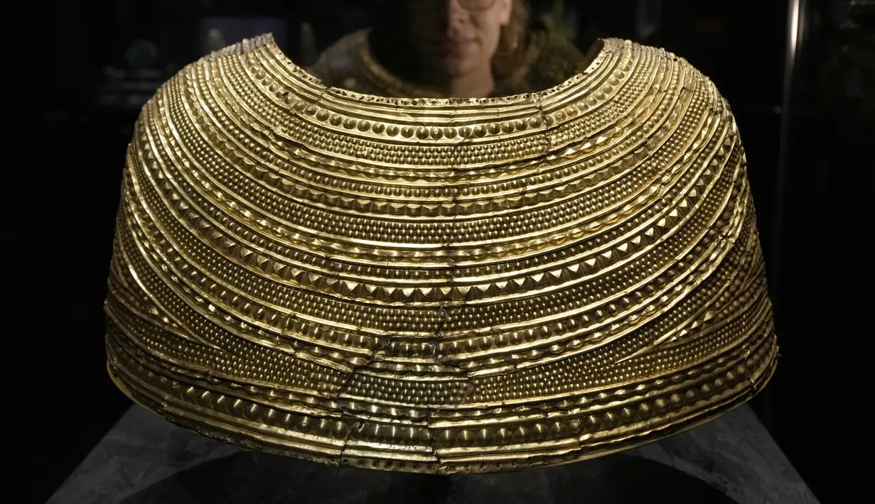 Seorang anggota staf berpose di sebelah jubah emas yang ditemukan di Mold in Wales, tertanggal 1600-1900 SM, dipajang di pameran The World of Stonehenge' di British Museum di London, Senin (14/2/2022). (AP Photo/Alastair Grant)