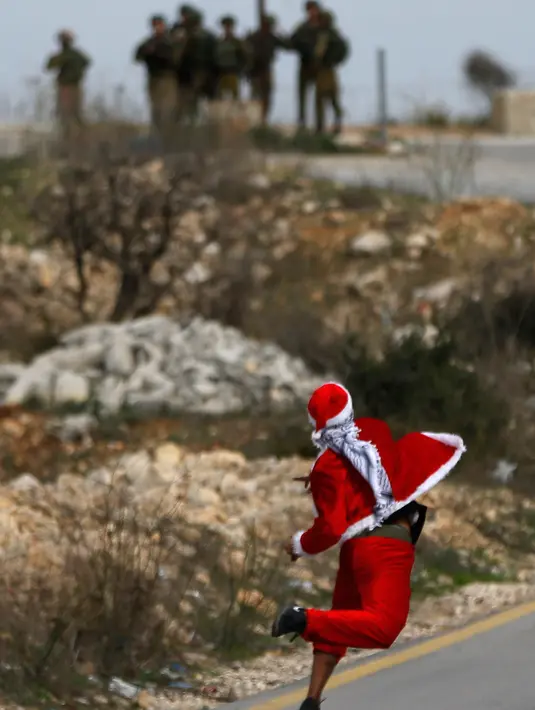 Seorang demonstran Palestina mengenakan kostum Santa Claus melempar batu ke arah pasukan keamanan Israel saat terjadi bentrokan di pos pemeriksaan Atarot di pinggiran utara Yerusalem (19/12). (AFP Photo/Abbas Momani)